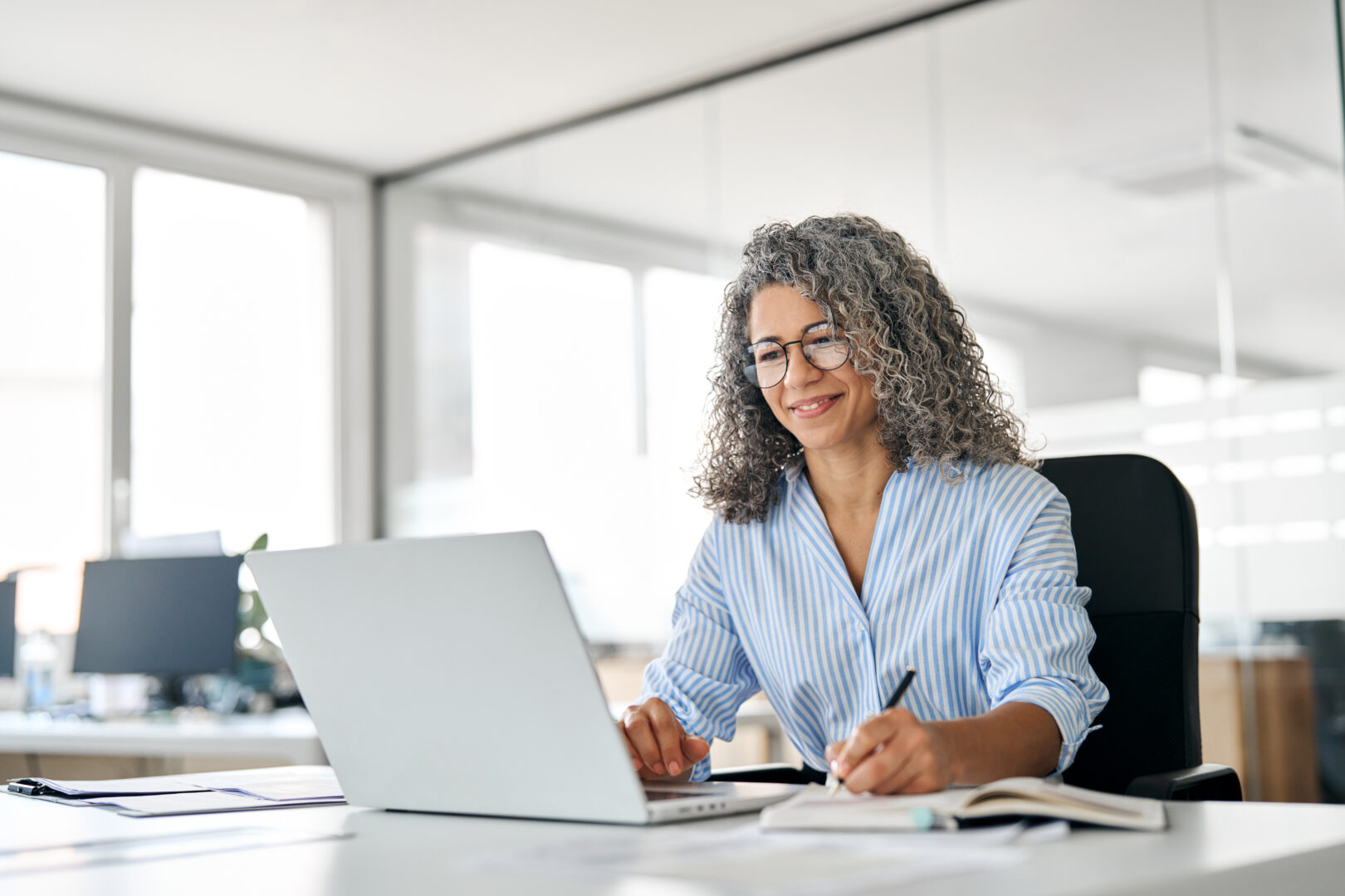 Busy mature business woman working in office using laptop writing notes.
